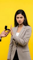 Young South Asian businesswoman in beige suit refusing interview microphone with stop gesture on bright yellow background for media concepts.