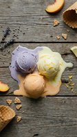 Three scoops of lavender, vanilla, and chocolate ice cream on rustic wooden surface with peach slices, waffle cone pieces, and dried lavender for dessert and food styling projects.
