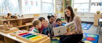 Female teacher reading storybook to diverse group of young children sitting on colorful classroom carpet during story time learning activity.