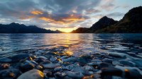 Dramatic mountain lake sunset with smooth stones underwater in foreground, golden light reflecting on rippling water surface against dramatic cloudy sky.