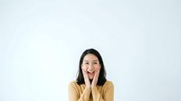 Young woman with long dark hair and South Asian ethnicity wearing a beige cardigan, resting her face on her hands with an engaged, cheerful expression against a white background.