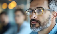 Close up portrait of mature Caucasian businessman with gray beard and glasses looking thoughtfully ahead against blurred office background with bokeh lights.