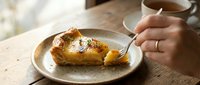 Woman enjoying slice of caramelized upside-down pineapple cake with fork on rustic ceramic plate. Cozy cafe atmosphere with warm lighting and tea cup.
