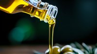 Golden cooking oil pouring from glass bottle in smooth stream against dark background with green bokeh lights for culinary and kitchen applications.