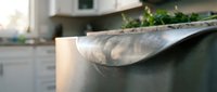 Close-up of a shiny stainless steel pot rim with steam rising in a modern kitchen, blurred background with white cabinets and fresh herbs.