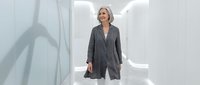 Professional middle-aged woman with blonde hair wearing gray blazer in modern white corridor with neon lighting. Corporate business environment.