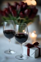Two glasses of red wine with wrapped gift box and dark red tulips in soft focus background, romantic evening setting with warm bokeh lights.