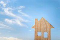Wooden house symbol against blue sky with white clouds representing home ownership, real estate investment, property market and housing dreams.