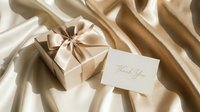 Elegant gift box with satin ribbon and thank you card on luxurious golden silk fabric, perfect for wedding, holiday, or celebration concepts.