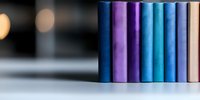 Row of hardcover books in blue and purple shades standing on white surface against blurred background with bokeh lights, creating minimalist composition.