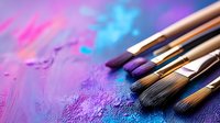 Artist paintbrushes on vibrant purple and blue textured background with scattered pigment powder, creating atmospheric artistic composition for creative projects.