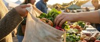 Two people exchanging fresh vegetables in reusable cloth bag at farmers market or outdoor produce stand during daytime shopping.