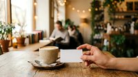 Cropped hand holding blank business card next to coffee cup on wooden table in modern cafe with blurred staff and holiday decorations in background.