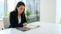 Professional businesswoman in dark suit reading documents at modern office desk with tablet and city view for corporate presentations and workplace content.