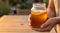 Unrecognizable person holding glass jar of golden honey outdoors on wooden table with blurred garden background, suitable for food and beverage marketing.