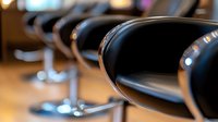 Row of empty black salon styling chairs with chrome bases in modern hair salon with blurred background, ready for clients.