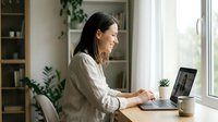 Woman with dark hair, South Asian ethnicity, wearing a beige linen shirt, sitting at a desk working on a laptop in a bright home office with plants and natural light from windows.
