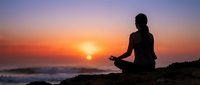 Woman silhouette meditating in lotus pose during sunset by ocean. Peaceful yoga practice on rocky coastline with golden sky and calm waves for wellness.