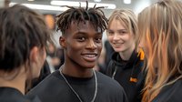 Diverse young people socializing at indoor event, focus on smiling African American male with dreadlocks wearing black t-shirt and silver chain necklace.