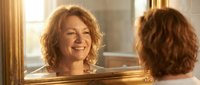 Middle-aged Caucasian woman with curly hair smiling at her reflection in ornate golden mirror, self-care and positive aging concept for wellness campaigns.