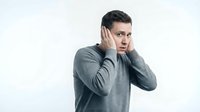 Young Caucasian man in gray sweater covering ears with hands showing shocked surprised expression isolated on white background for design use.