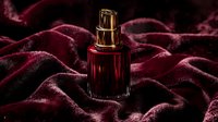 Red glass perfume bottle with gold cap on luxurious burgundy velvet fabric background with soft folds creating elegant shadows and reflections.