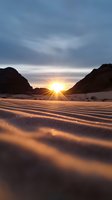 Dramatic sunset over desert sand dunes with rippled patterns casting long shadows, silhouetted mountains frame golden sunburst through valley gap in moody sky.