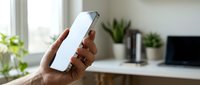 Hand holding a smartphone with blank white screen mockup in a bright home office, ready for app or website display.