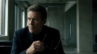 Middle-aged man with gray hair and facial stubble, wearing dark business suit, holding pen thoughtfully in modern office corridor with concrete walls and natural light from windows.