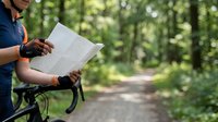 Cyclist in dark blue shirt holding map while standing on forest trail with blurred green trees and pathway in background, planning route during outdoor biking adventure.