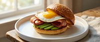 Gourmet breakfast sandwich with fried egg, bacon and avocado on sesame seed bun served on white plate in bright kitchen setting for food photography.