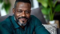 Confident mature African American businessman with gray beard wearing teal sweater smiling warmly at camera, indoor office setting with natural light and plants.
