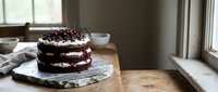 Homemade chocolate layer cake with cream frosting and berry topping on marble board near kitchen window for baking and dessert photography.