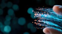 Glowing blue fiber optic strands with orange light particles streaming between fingers against dark background, representing digital transformation and data flow.