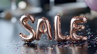 Rose gold foil balloon letters spelling SALE with scattered sparkly confetti on reflective surface against blurred background for retail promotion and marketing.
