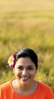 Young woman with dark hair and Southeast Asian ethnicity wearing bright orange shirt, smiling at camera with pink and yellow flower in hair against blurred green field background.