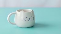 White ceramic cat-shaped mug with cute face and ears on mint green background. Adorable kitty coffee cup for pet lovers and kawaii style enthusiasts.