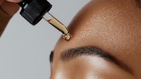 Close-up of African American woman applying facial serum with dropper, showing skincare routine and glowing, hydrated skin texture on forehead area.