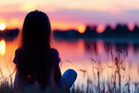 Young woman silhouette watching sunset over lake, sitting among tall grass with orange and purple sky reflecting on calm water surface. Peaceful solitary moment.