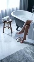 Woman's bare legs beside a vintage clawfoot bathtub in a luxury marble bathroom with towel, slippers, and wooden stool for spa self-care concept.