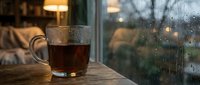 Steaming hot tea cup on wooden table by rainy window with cozy living room background. Perfect for relaxation and comfort themes.