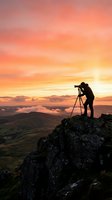 Photographer silhouette capturing sunrise from mountain peak with tripod camera equipment against dramatic orange pink sky and rolling hills landscape.