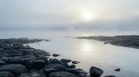 Serene coastal landscape with large boulders lining rocky shoreline at dawn, calm waters reflecting soft sunlight through overcast sky, ideal for travel and nature backgrounds.