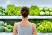 Young woman in gray tank top looking at fresh vegetables and greens on supermarket shelves, back view. Healthy shopping and nutrition concept.