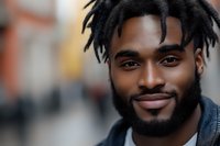 Young African American man with dreadlocks and beard smiling confidently at camera, wearing denim jacket against blurred urban background. Natural daylight portrait.