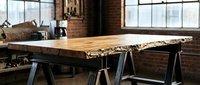Live edge wood table with natural bark on metal sawhorse legs in industrial loft workspace with brick walls and large windows for modern furniture design.