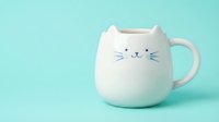 Adorable white ceramic cat mug with cute face design on mint green background. Kawaii style coffee cup for morning beverages and cozy moments.