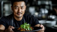 Professional Asian male chef presenting gourmet dish with fresh herbs in modern restaurant kitchen for culinary marketing and food service industry.