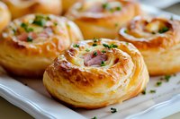 Flaky puff pastry pinwheels filled with creamy cheese and ham, garnished with fresh chopped herbs on white ceramic plate, close-up view of golden baked appetizers.