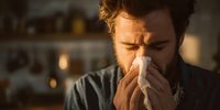 Man with beard blowing nose into tissue at home during illness, warm lighting creates atmospheric mood for health concept.